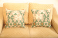Green and beige geometric wool square cushion set of two with cream and rust accents displayed on a beige sofa.